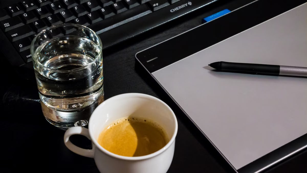 Office desk setup with a coffee cup, glass of water, tablet, pen, and keyboard.